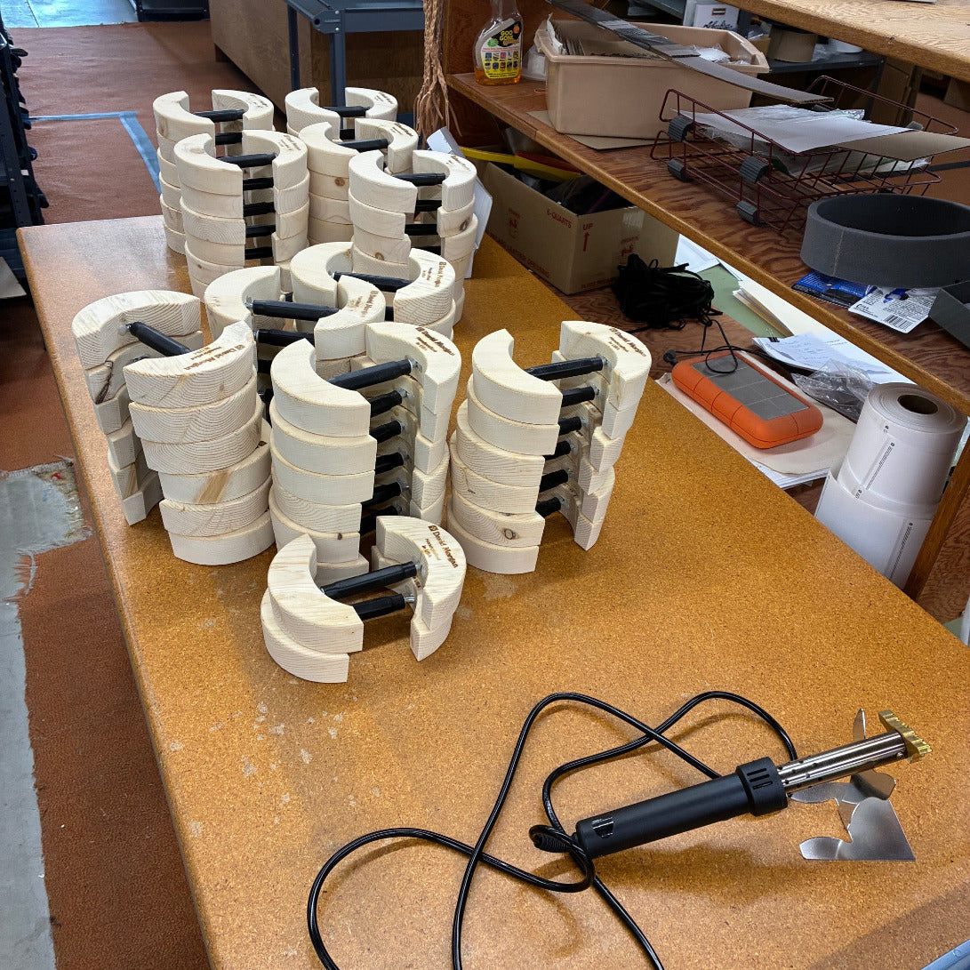 Multiple white curved plastic or wood clamping pieces with black handles are arranged on a workbench, alongside a black soldering iron with a brass tip. The setting is a workshop with shelves and tools in the background.